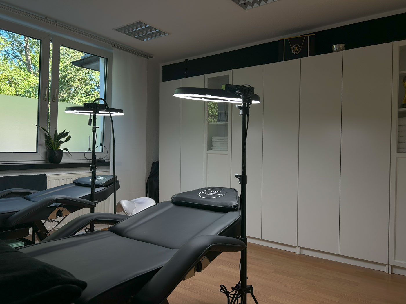 Leerer moderner Behandlungsraum mit zwei schwarzen Liegen, hellen Schränken und großen Fenstern.